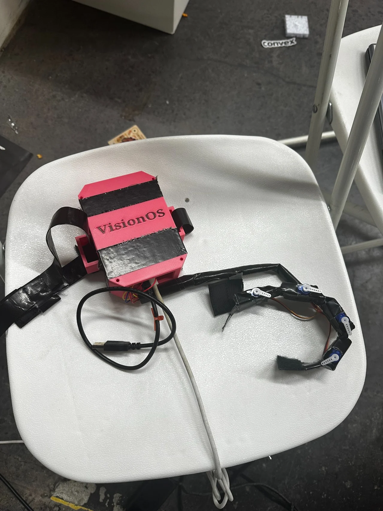 VisionOS prototype: a pink enclosure connected by cables to a curved wearable assembly with actuators, resting on a white chair.